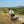 A cyclist wearing a black helmet and light beige shirt rides a bicycle on a gravel path through a scenic, grassy mountain landscape under a clear blue sky. A Wholegrain Cycles Carrie rack and bag are attached to the handlebars.
