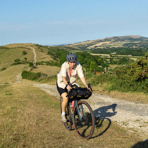 A cyclist wearing a black helmet and light beige shirt rides a bicycle on a gravel path through a scenic, grassy mountain landscape under a clear blue sky. A Wholegrain Cycles Carrie rack and bag are attached to the handlebars.