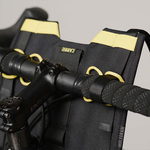 Close-up of a black bicycle handlebar bag with bright yellow accents and loops, mounted over black handlebars. The brand name 'Carrie' is visible on a small black label.