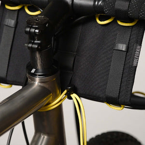 Close-up of a black WHOLEGRAIN bicycle handlebar bag mounted on a dark metallic road bike frame, secured with yellow bungee cords.