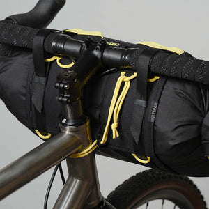 A close-up of a black Wholegrain Cycles Carrie bicycle handlebar bag mounted over the handlebars, secured with black straps and yellow drawstrings, on a grey bicycle against a light grey background.