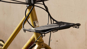 A close-up of a yellow bicycle frame with a Wholegrain Cycles Jack Black bike rack mounted on the seat tube, shown against a plain beige wall.