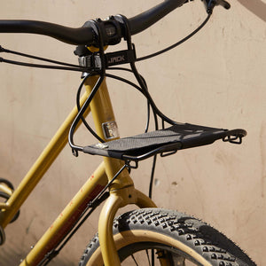 A close-up of a yellow bicycle frame with a Wholegrain Cycles Jack Black bike rack mounted on the seat tube, shown against a plain beige wall.