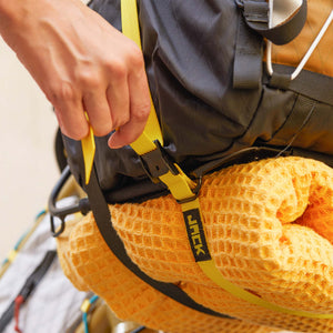 Close-up of a person's hand fastening a yellow Wholegrain Cycles Stiff strap around a black backpack with an orange sleeping pad underneath.