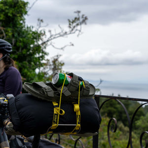 A black bicycle pannier bag loaded with olive green sandals and a green can, secured with Wholegrain Cycles yellow Stiff straps, set against an outdoor scenic background with a cyclist partially visible on the left.