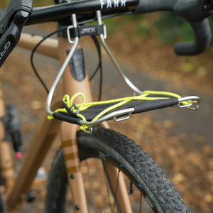 A close-up of a Kinesis Tripster AT bicycle with a Wholegrain Cycles Jack Classic Rack mounted on the handlebars using silver metal clips and bright yellow-green bungee cords, photographed outdoors on a blurred path.