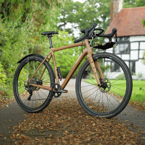 A Kinesis Tripster AT gravel bicycle with a natural wood-look frame and black components including a Wholegrain Cycles Jack Classic Rack , parked on a leaf-strewn path with a traditional English cottage in the background.